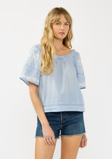 [Color: Light Blue] A front facing image of a blonde model wearing a light blue embroidered top with short sleeves and a scoop neckline with ruffle trim.