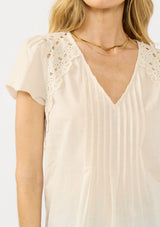 [Color: Natural] A detail image of a blonde model wearing a lightweight beige top with embroidered cutouts, pintucks, and a v neckline.