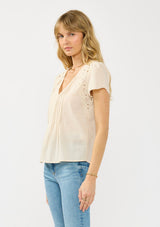 [Color: Natural] A side facing image of a blonde model wearing a lightweight beige top with embroidered cutouts, pintucks, and a v neckline.