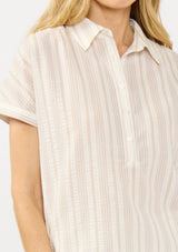 [Color: Sand/Off White] A detail image of a blonde model wearing a cotton striped shirt with a puckered texture, collar neck, button front, and short sleeves. Styled with matching shorts. 
