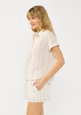 [Color: Sand/Off White] A side facing image of a blonde model wearing a cotton striped shirt with a puckered texture, collar neck, button front, and short sleeves. Styled with matching shorts. 