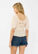[Color: Natural] A back facing image of a blonde model wearing a beige puff sleeve top with a square neckline, lace front, pintuck details, and a tie back detail. 