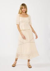 [Color: Natural] A front facing image of a blonde model wearing a beige puff sleeve top with a square neckline, lace front, pintuck details, and a tie back detail. Styled with a matching beige lace maxi skirt.