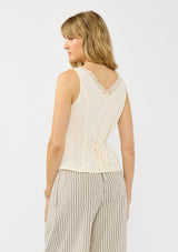 [Color: Tea] A back facing image of a blonde model wearing an off white tank top with lace trims, a button fort, inverted pleats, and a tie back waist detail. Styled with beige striped bottoms. 