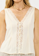 [Color: Tea] A close up image of a blonde model wearing an off white tank top with lace trims, a button fort, inverted pleats, and a tie back waist detail. Styled with beige striped bottoms.  