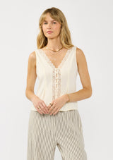 [Color: Tea] A front facing image of a blonde model wearing an off white tank top with lace trims, a button fort, inverted pleats, and a tie back waist detail. Styled with beige striped bottoms. 