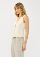[Color: Tea] A side facing image of a blonde model wearing an off white tank top with lace trims, a button fort, inverted pleats, and a tie back waist detail. Styled with beige striped bottoms. 