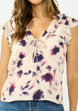 [Color: Peach/Plum] A detail image of a brunette model wearing a pink and purple floral top with flutter sleeves, a v neckline with ruffle trim, and a tie front detail. 