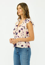 [Color: Peach/Plum] A side facing image of a brunette model wearing a pink and purple floral top with flutter sleeves, a v neckline with ruffle trim, and a tie front detail. 