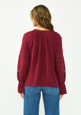 [Color: Merlot] A back facing image of a brunette model wearing a burgundy bohemian blouse with a button front, v neckline, lace trims, and pintuck details. 