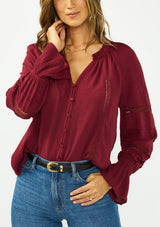 [Color: Merlot] A detail image of a brunette model wearing a burgundy bohemian blouse with a button front, v neckline, lace trims, and pintuck details. Paired with a light wash denim and a black belt. 