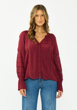 [Color: Merlot] A front facing image of a brunette model wearing a burgundy bohemian blouse with a button front, v neckline, lace trims, and pintuck details. 
