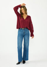 [Color: Merlot] A front facing image of a brunette model wearing a burgundy bohemian blouse with a button front, v neckline, lace trims, and pintuck details. Paired with a light blue denim, a black belt, and black boots. 