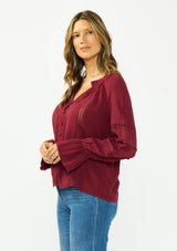[Color: Merlot] A side facing image of a brunette model wearing a burgundy bohemian blouse with a button front, v neckline, lace trims, and pintuck details. 