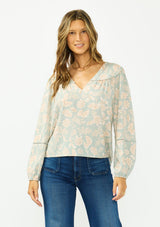 [Color: Dusty Sage/Natural] A front facing image of a brunette model wearing a mint floral blouse with a v neckline, front yoke, and long sleeves. Paired with a dark wash denim with patch pockets. 
