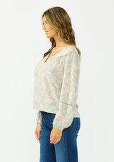 [Color: Dusty Sage/Natural] A side facing image of a brunette model wearing a mint floral blouse with a v neckline, front yoke, and long sleeves. Paired with a dark wash denim with patch pockets. 