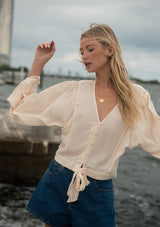 [Color: Tea Dye] A front facing image of a blonde model at the beach wearing a light beige blouse with wide long sleeves, a v neckline, button front, and a tie front detail. 