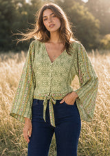 [Color: Olive/Rose] A front facing image of a brunette model in the fields wearing a green bohemian floral top with wide sleeves, a v neckline,  a button front, and a tie front detail. Styles with a dark wash denim with patch pockets. 