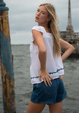 [Color: White/Blue] A back facing image of a blonde model near water wearing a white flutter sleeve top with blue floral embroidery, a button front, and pintucks. 