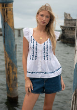 [Color: White/Blue] A front facing image of a blonde model near water wearing a white flutter sleeve top with blue floral embroidery, a button front, and pintucks. 