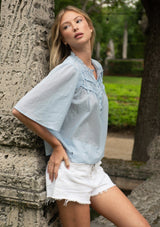 [Color: Blue] Woman in light blue blouse and white shorts leaning against a stone wall in an outdoor setting.