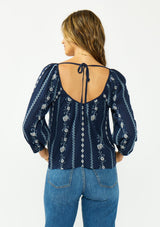 [Color: Navy/Blue] A back facing image of a brunette model wearing a blue floral embroidered blouse with a wide neckline, three quarter length sleeves, and a tie back detail. 