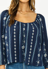 [Color: Navy/Blue] A detail image of a brunette model wearing a blue floral embroidered blouse with a wide neckline, three quarter length sleeves, and a tie back detail. 
