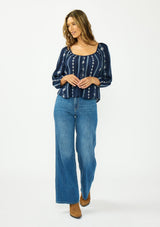 [Color: Navy/Blue] A front facing image of a brunette model wearing a blue floral embroidered blouse with a wide neckline, three quarter length sleeves, and a tie back detail. Styles with blue denim jeans and brown boots. 