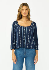 [Color: Navy/Blue] A front facing image of a brunette model wearing a blue floral embroidered blouse with a wide neckline, three quarter length sleeves, and a tie back detail. 
