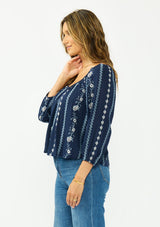 [Color: Navy/Blue] A side facing image of a brunette model wearing a blue floral embroidered blouse with a wide neckline, three quarter length sleeves, and a tie back detail. 
