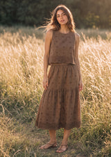 [Color: Brown] A front facing image of a brunette model in the fields wearing a brown embroidered tank top and midi skirt set.