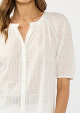[Color: Natural/Cream] A detail image of a brunette model wearing a white embroidered puff sleeve top with a button front. 