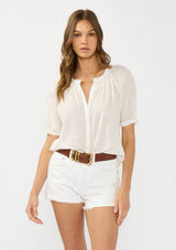[Color: Natural/Cream] A front facing image of a brunette model wearing a white embroidered puff sleeve top with a button front. Paired with white denim shorts and a brown belt. 