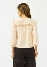 [Color: Shell] A back facing image of a brunette model wearing a nude lace top with long sleeves, a v neckline, and a button front. Paired with a dark brown denim. 