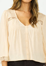 [Color: Shell] A detail image of a brunette model wearing a nude lace top with long sleeves, a v neckline, and a button front. Paired with a dark brown denim. 