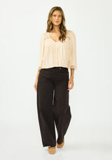 [Color: Shell] A front facing image of a brunette model wearing a nude lace top with long sleeves, a v neckline, and a button front. Paired with a dark brown denim. 
