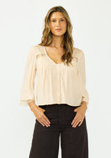[Color: Shell] A front facing image of a brunette model wearing a nude lace top with long sleeves, a v neckline, and a button front. Paired with a dark brown denim. 