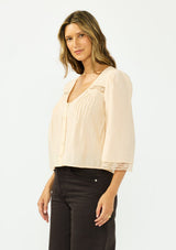 [Color: Shell] A side facing image of a brunette model wearing a nude lace top with long sleeves, a v neckline, and a button front. Paired with a dark brown denim. 