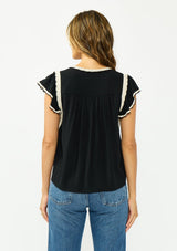 [Color: Black/Natural] A back facing image of a brunette model wearing a black flutter sleeve top with cream crochet trims, a v-neckline, and pintuck details. 