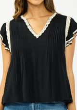 [Color: Black/Natural] A detail image of a brunette model wearing a black flutter sleeve top with cream crochet trims, a v-neckline, and pintuck details. 