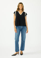 [Color: Black/Natural] A front facing image of a brunette model wearing a black flutter sleeve top with cream crochet trims, a v-neckline, and pintuck details. Paired with blue denim and black closed toe heels. 