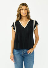 [Color: Black/Natural] A front facing image of a brunette model wearing a black flutter sleeve top with cream crochet trims, a v-neckline, and pintuck details. 