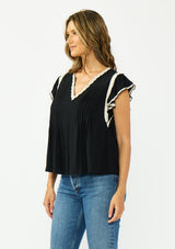 [Color: Black/Natural] A side facing image of a brunette model wearing a black flutter sleeve top with cream crochet trims, a v-neckline, and pintuck details. 