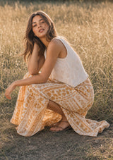 [Color: Cream/Natural] A sitting image of a brunette model in a field wearing a beige eyelet tank top with a square neckline, tie detail, and a scalloped trim. Styles with a yellow bohemian skirt. 