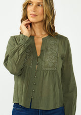 [Color: Olive] A front facing image of a brunette model wearing an olive green embroidered blouse with a button front and a button cuff closure with ruffle trim.