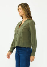 [Color: Olive] A side facing image of a brunette model wearing an olive green embroidered blouse with a button front and a button cuff closure with ruffle trim. Paired with a dark wash denim.