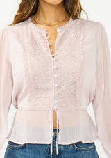 [Color: Lilac] A detail image of a brunette model wearing a sheer lilac top with a front lace accent, button front, long sleeves, and a tie front waist detail. 