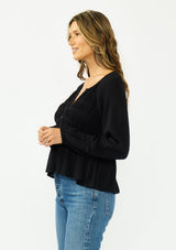 [Color: Black] A side facing image of a brunette model wearing a black long sleeve top with a shirred bust area. 