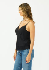 [Color: Black] A side facing image image of a brunette model wearing a black lace cami with spaghetti straps. Styled with dark wash denim. 