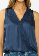 [Color: Midnight] A detail image of a brunette model wearing a blue satin sleeveless top with an embroidered trim, elastic hem, and a surplice front. 